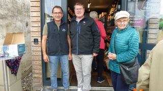 Three people stand outside a shop. A man on the left wears a vest and glasses. The central man wears a jacket. A woman on the right wears a hat and a jacket. Shelves and products are behind them.