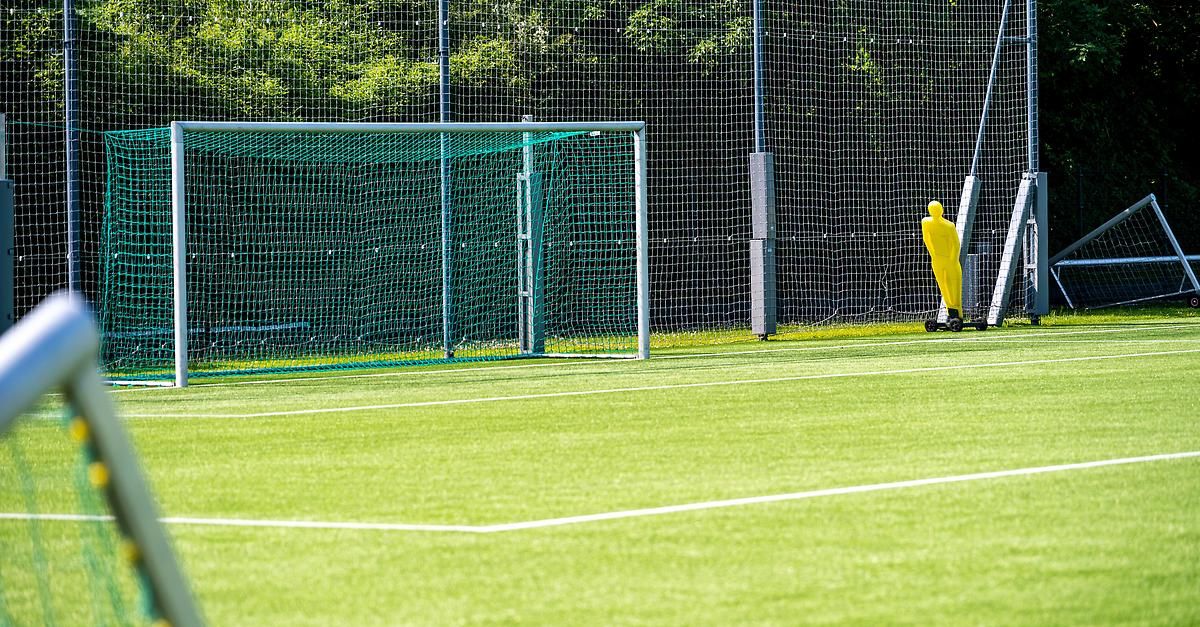 An empty soccer field with a goal and a yellow mannequin on the right. The field is surrounded by a net fence.