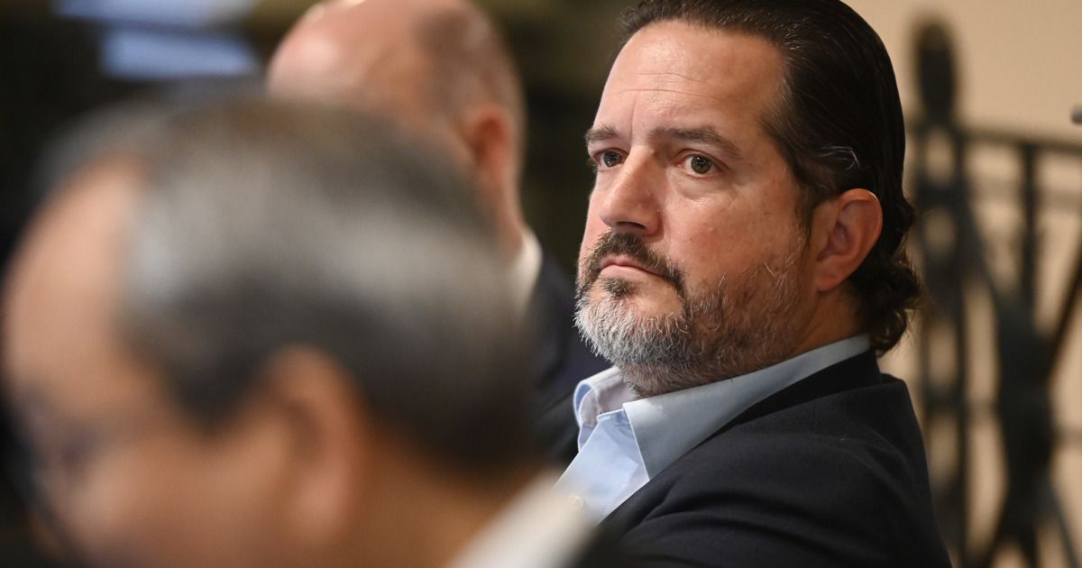 A man with a beard is sitting in a meeting, looking focused. He wears a blue shirt under a black jacket. Another person's head is blurred in the foreground.