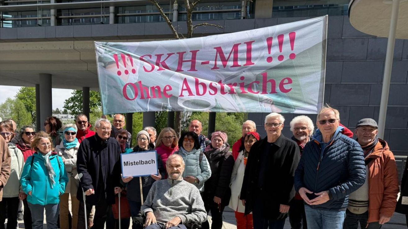 A group of elderly people pose in front of a banner with the text '!!! SKH-MI!!! Ohne Abstriche'. They are smiling and appear to be protesting.