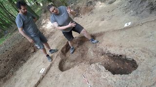 Two individuals stand on a dirt site, one gesturing with her hands. A large, excavated hole is present, with a white label nearby. The site is surrounded by trees.