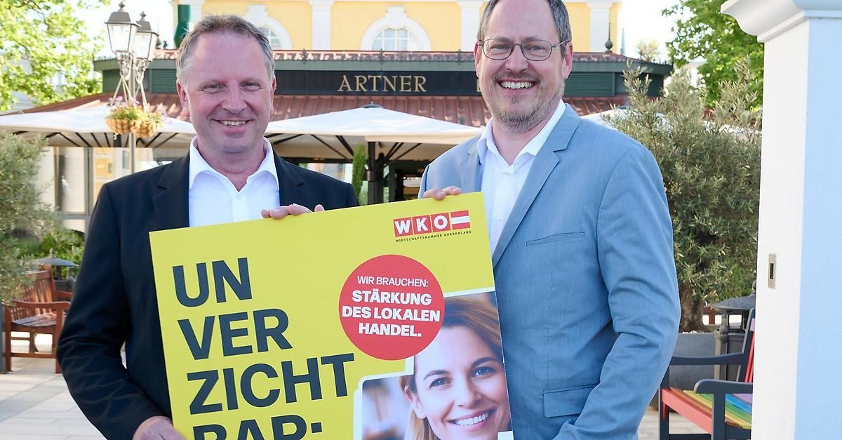 Zwei Männer stehen vor einem Restaurant mit einem Schild, auf dem 'UN VERSICHT BAR' steht und einem Logo. Der Mann auf der rechten Seite lächelt und hält ein Banner.