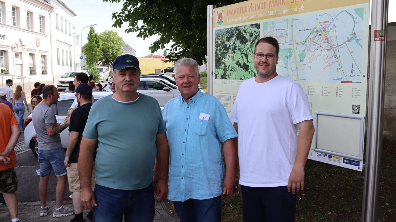Three men stand outside with a sign in the background, one with a green shirt, another with a blue shirt, and the third with a white shirt.
