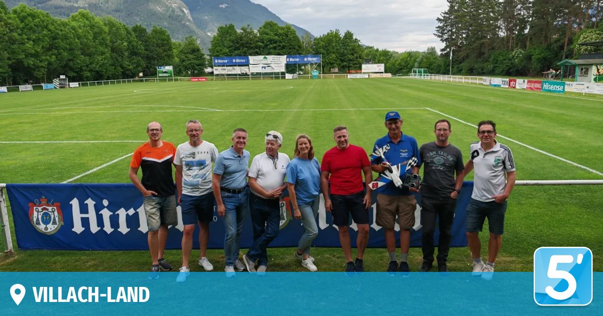 Nine adults pose for a photo at a soccer field. One man holds a toy car. Behind them are banners and trees.