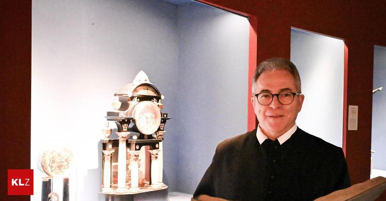 A man with glasses stands next to an antique clock, smiling and posing for a photo.