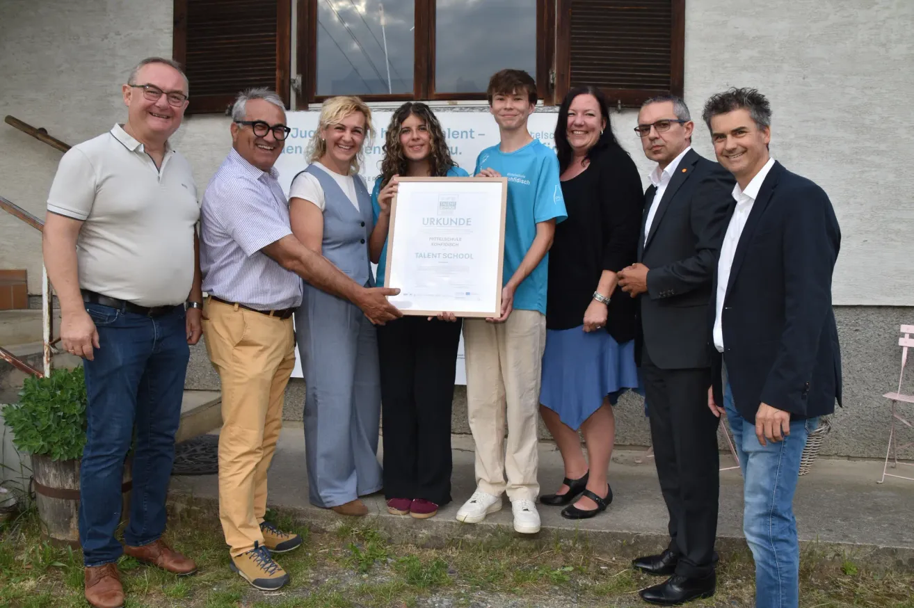 A group of people, including a man holding a framed certificate, stand on a concrete platform. The man in the blue shirt holds the frame with both hands.