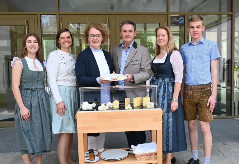 A group of people are standing next to a table with cheese and plates. The man in the middle holds a plate with cheese. Behind them is a building with glass doors.