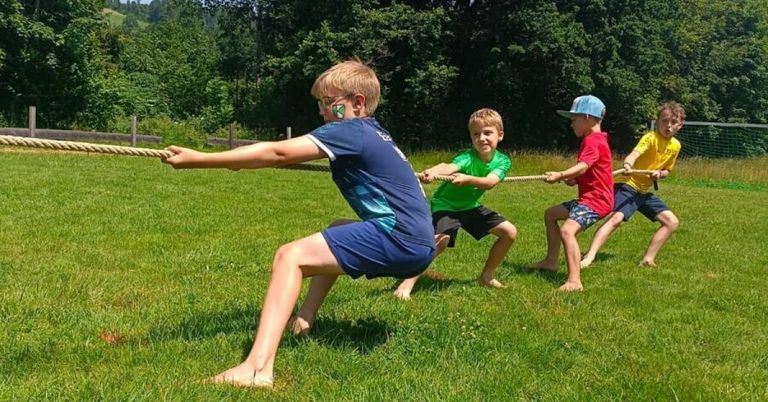 Drei Jungen ziehen an einem Seil auf einer Grasfläche mit Bäumen im Hintergrund.
