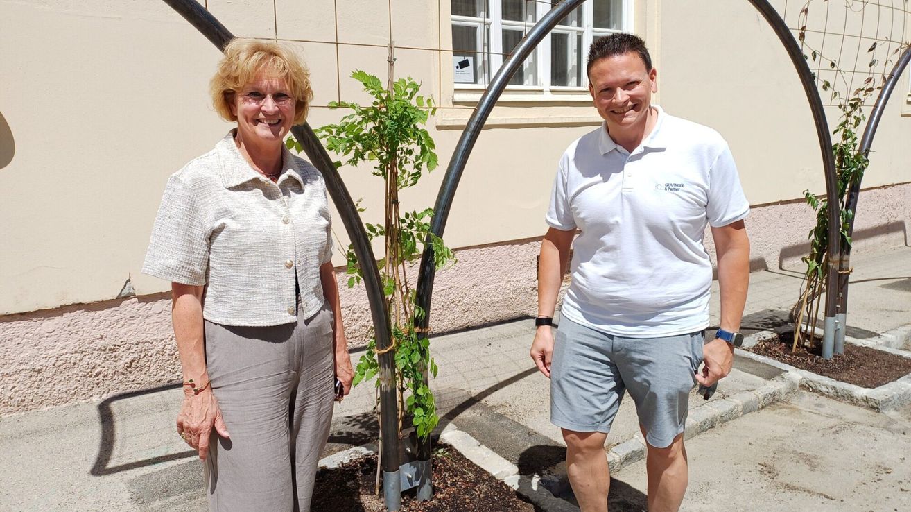 A woman and a man are standing next to a young tree on a sunny day. They are smiling and seem to be posing for a photo. The man is wearing a white polo shirt and the woman is wearing a white blouse.
