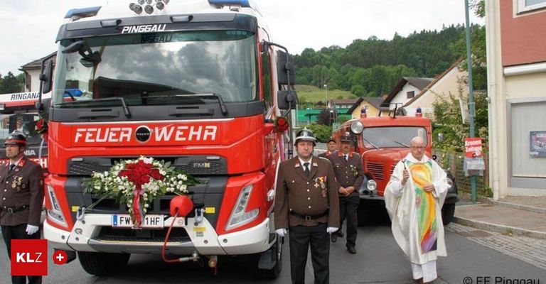 Ein Feuerwehrwagen mit einem Kranz steht in einem Dorf. Ein Mann in Uniform und ein Priester stehen neben ihm. Ein weiterer Feuerwehrwagen ist im Hintergrund.