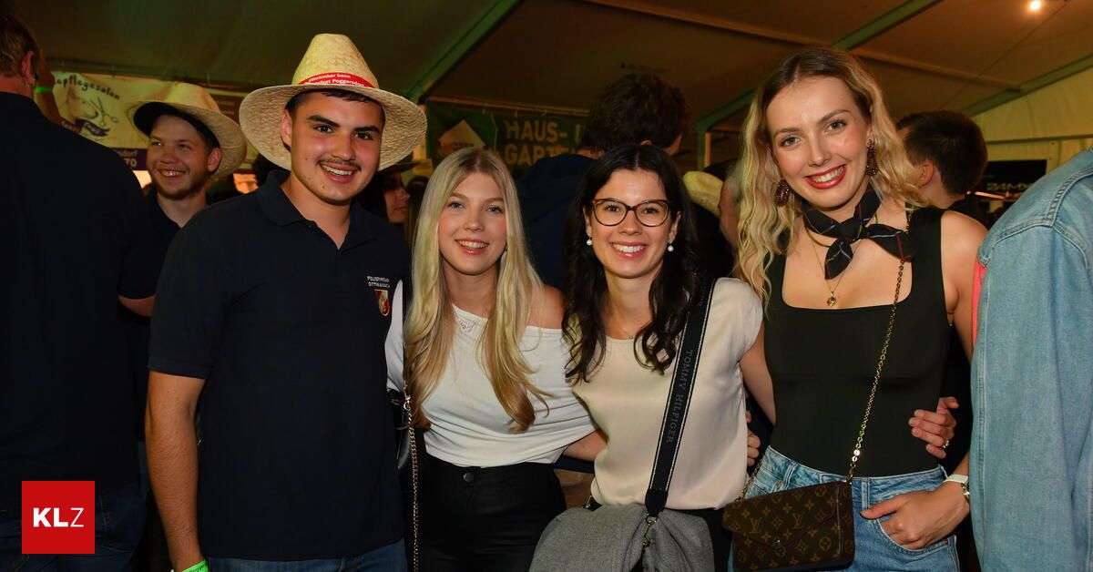 Vier junge Menschen posieren für ein Foto bei einem Event. Ein Mann und zwei Frauen lächeln. Die Frau mit Brille hält eine graue Tasche. Eine andere Frau hat eine Louis Vuitton-Handtasche. Dahinter sind noch mehr Menschen.
