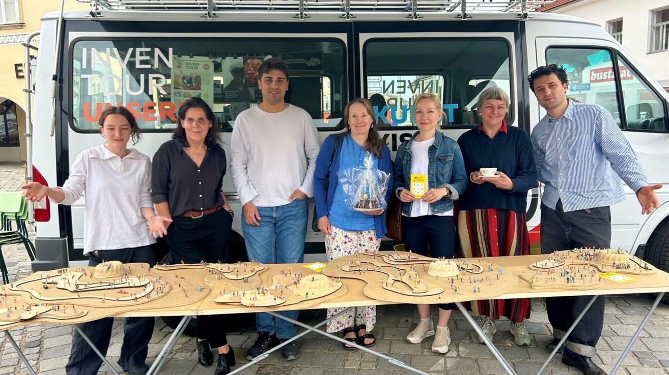 Five people stand behind a table with a model of a city. One woman holds a gift bag and a yellow card. Another holds a cup. The others smile. A tram is behind them.