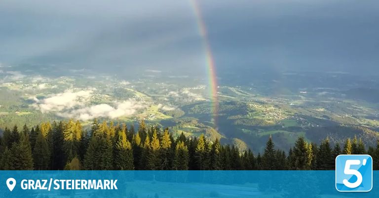 A panoramic view of a mountainous landscape with a rainbow in the sky and pine trees covering the foreground.