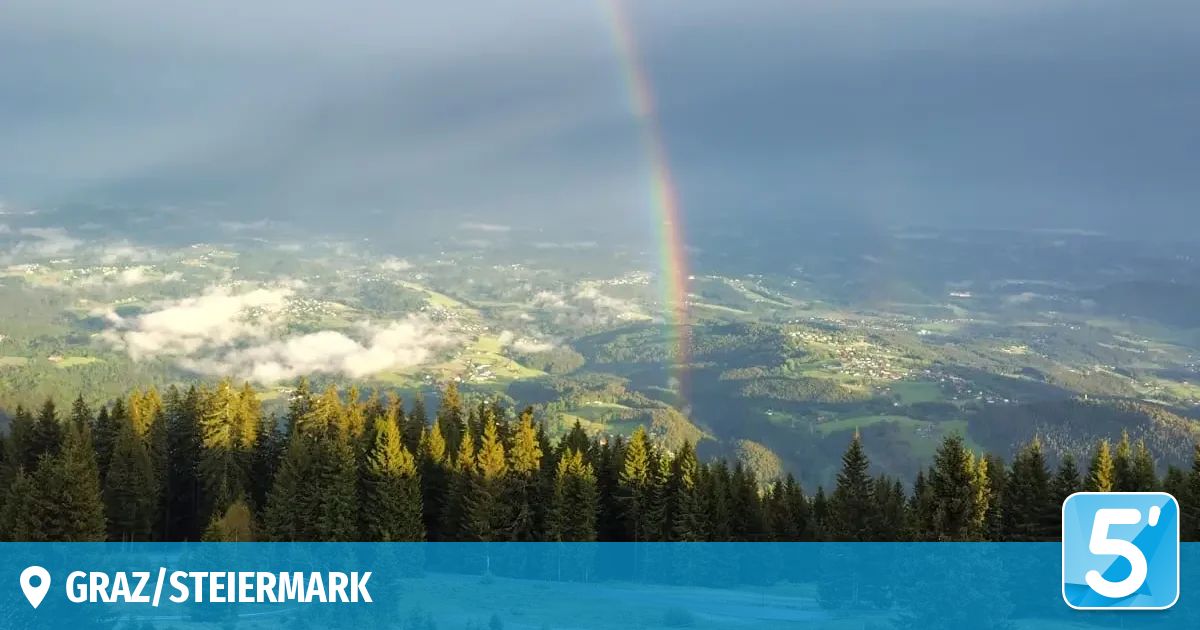A panoramic view of a mountainous landscape with a rainbow in the sky and pine trees covering the foreground.