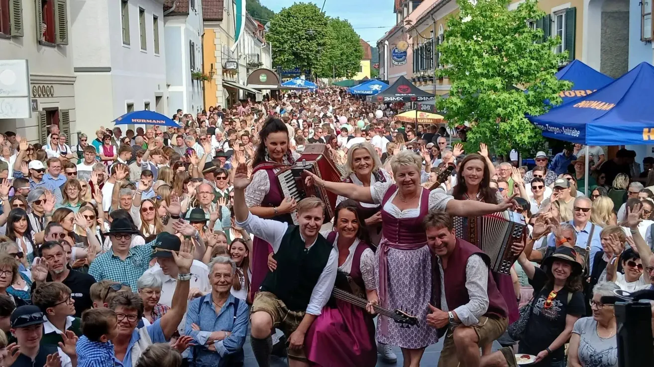 A large crowd of people in traditional Bavarian dress are performing on a street, with one person playing the accordion and another playing the guitar.