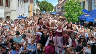 A large crowd of people in traditional Bavarian dress are performing on a street, with one person playing the accordion and another playing the guitar.