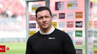 A man in a black sweater with a sky sport badge stands in front of a wall with many advertisements.