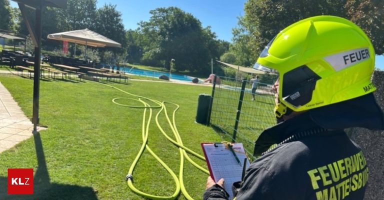 Ein Feuerwehrmann überprüft eine Checkliste in der Nähe eines Pools, mit einem grünen Schlauch auf dem Rasen. Bäume und ein Pavillon sind im Hintergrund.