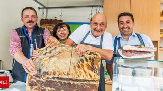 Four smiling people stand behind a large piece of meat. The man on the right holds a slice of meat. Behind them, there is a wooden shelf with more meat hanging.