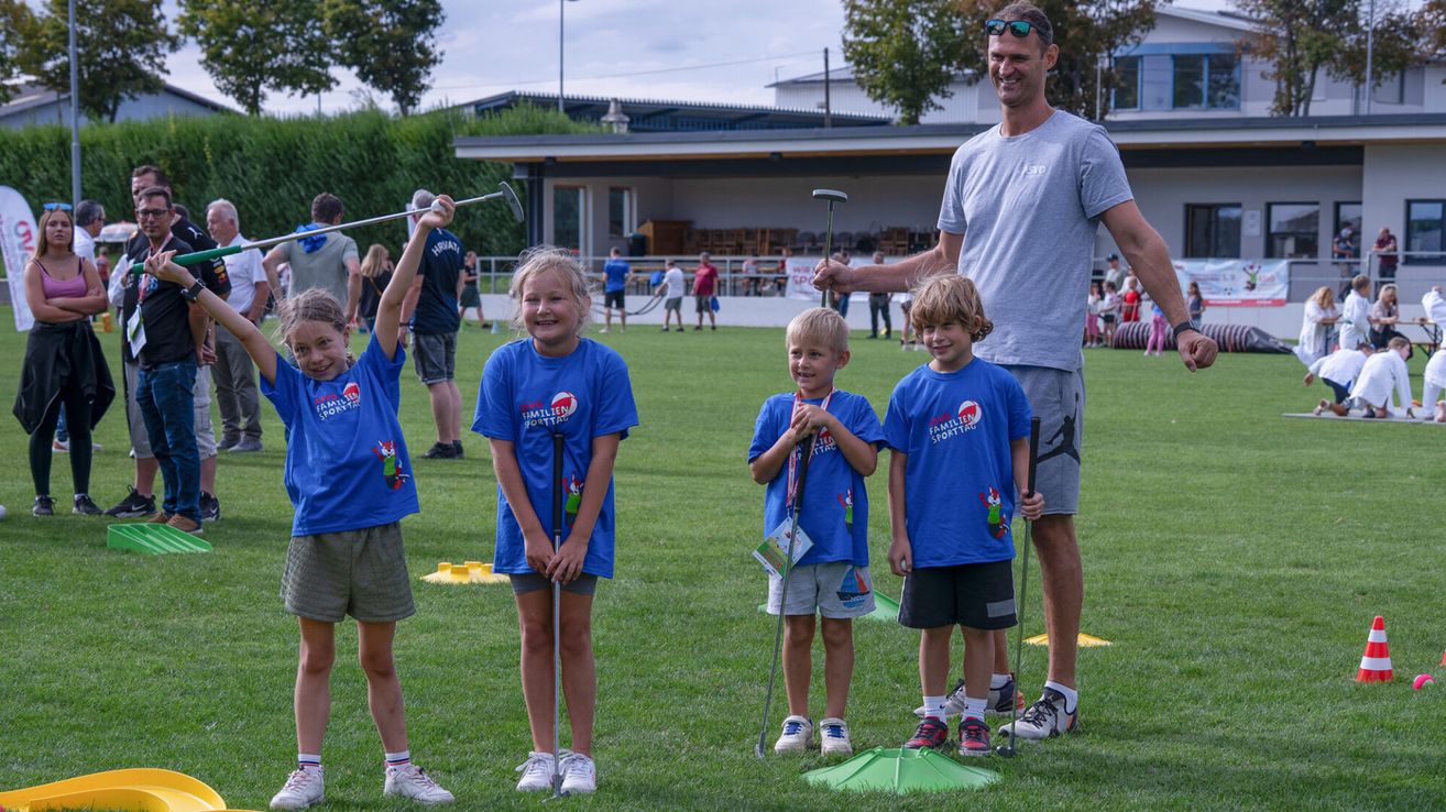 Ein Mann und drei Kinder halten Golfschläger auf einem Feld und lächeln. Die Kinder tragen passende Shirts und der Mann trägt eine Sonnenbrille. Hinter ihnen steht ein Gebäude und mehrere Menschen.