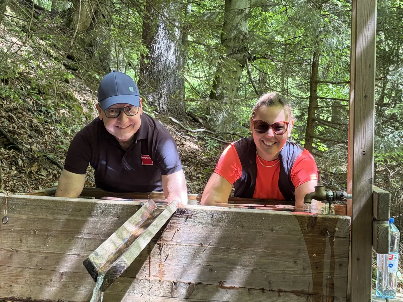 Two people are smiling at the camera in front of a wooden structure with water flowing through it, surrounded by trees.