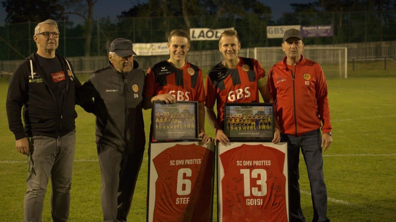 Vier Männer stehen auf einem Fußballfeld und halten gerahmte Fotos. Zwei tragen rote Uniformen mit den Nummern 6 und 13, einer trägt eine graue Jacke. Sie lächeln und schauen in die Kamera.