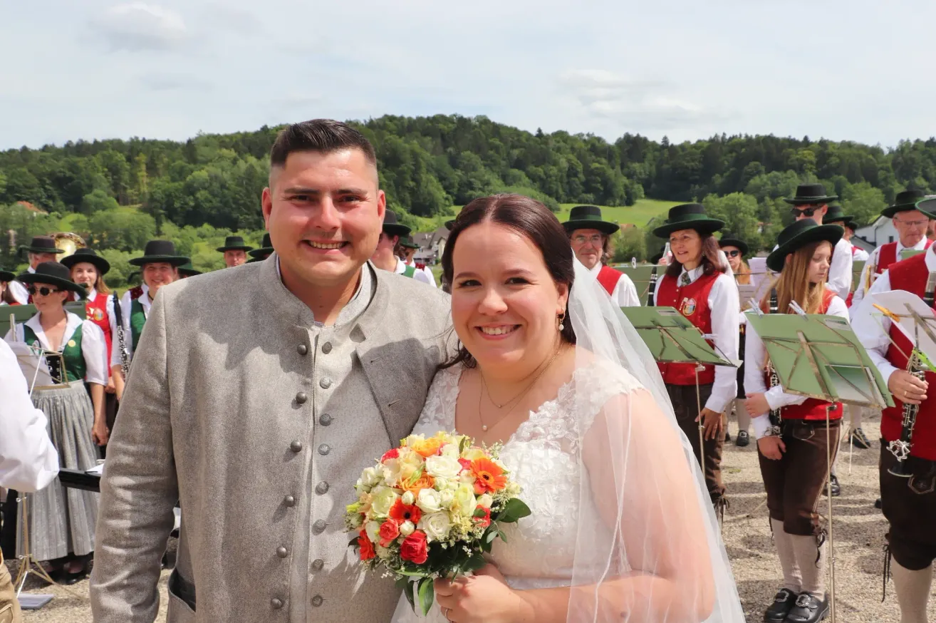 A smiling couple stands together in a garden with a band in the background, holding a bouquet of flowers. The groom wears a traditional suit, and the bride wears a white dress.