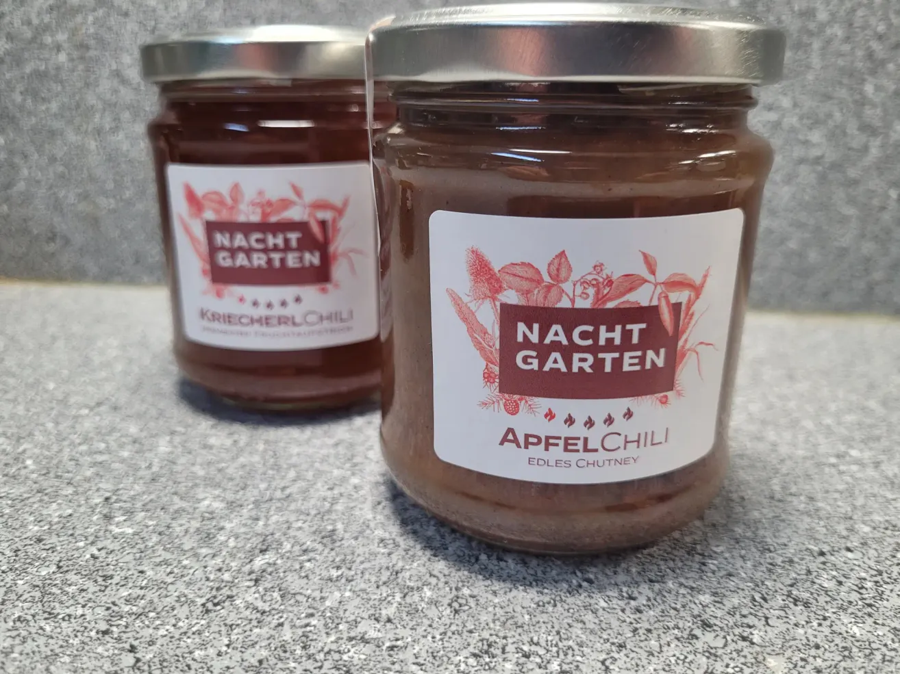 Two jars of Nacht Garten ApfelChili on a countertop. The front jar has a brown label with a chili pepper and the text Nacht Garten ApfelChili.