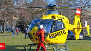 A rescue helicopter is parked on grass. A man in red is walking towards it. A police officer is standing behind it. Trees and a car are in the background.