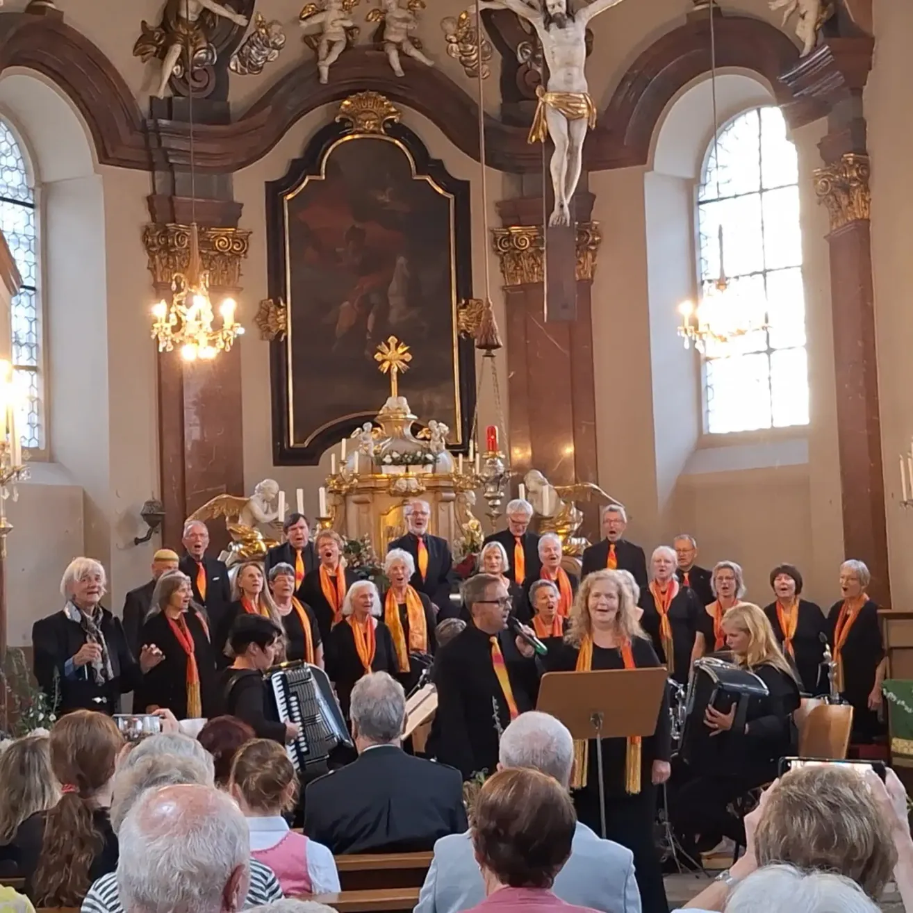 A choir of women and men wearing orange and black scarves performs in a church, standing in front of a golden altar with candles and a cross. A crowd of people is seated in front of them, and the room is illuminated by chandeliers.