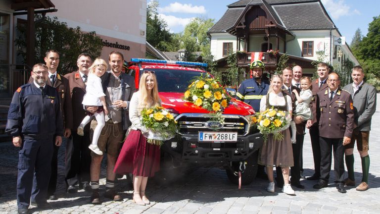 Eine Gruppe von Menschen in traditioneller deutscher Kleidung steht vor einem mit Blumen geschmückten Feuerwehrauto auf einer Kopfsteinpflasterstraße.