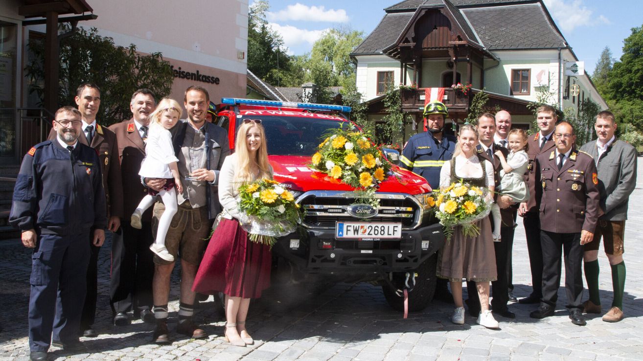 Eine Gruppe von Menschen in traditioneller deutscher Kleidung steht vor einem mit Blumen geschmückten Feuerwehrauto auf einer Kopfsteinpflasterstraße.
