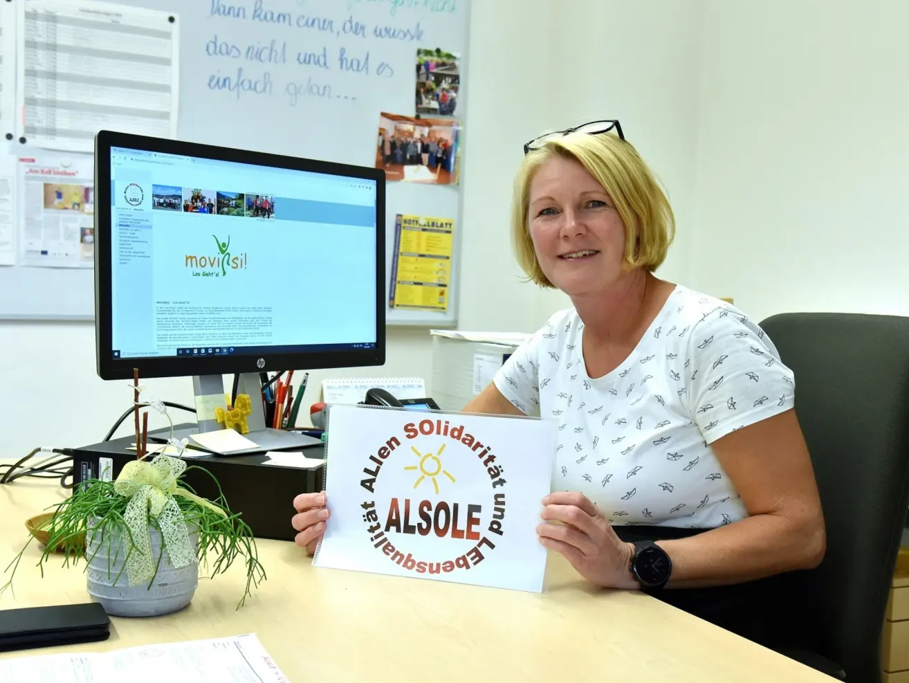 A woman sits at a desk with a computer, smiling. She holds a sign that reads 'Allen Solidarität und AlsOLE'. There is a potted plant, pens, and a monitor in the background.