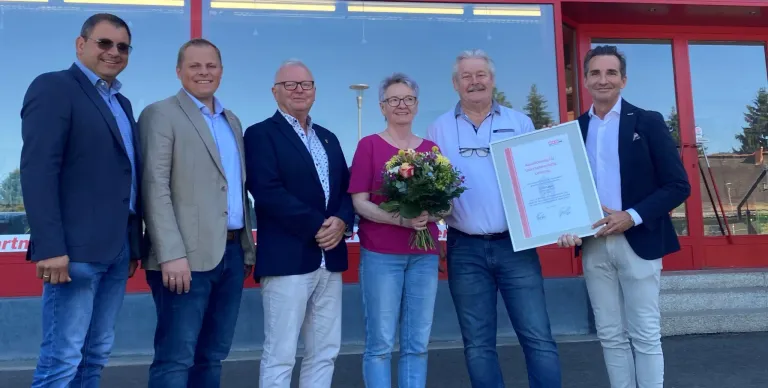 Four people stand in front of a building with flowers. A man on the left smiles, while the woman holds flowers. Another man holds a certificate. The glass wall reflects trees.