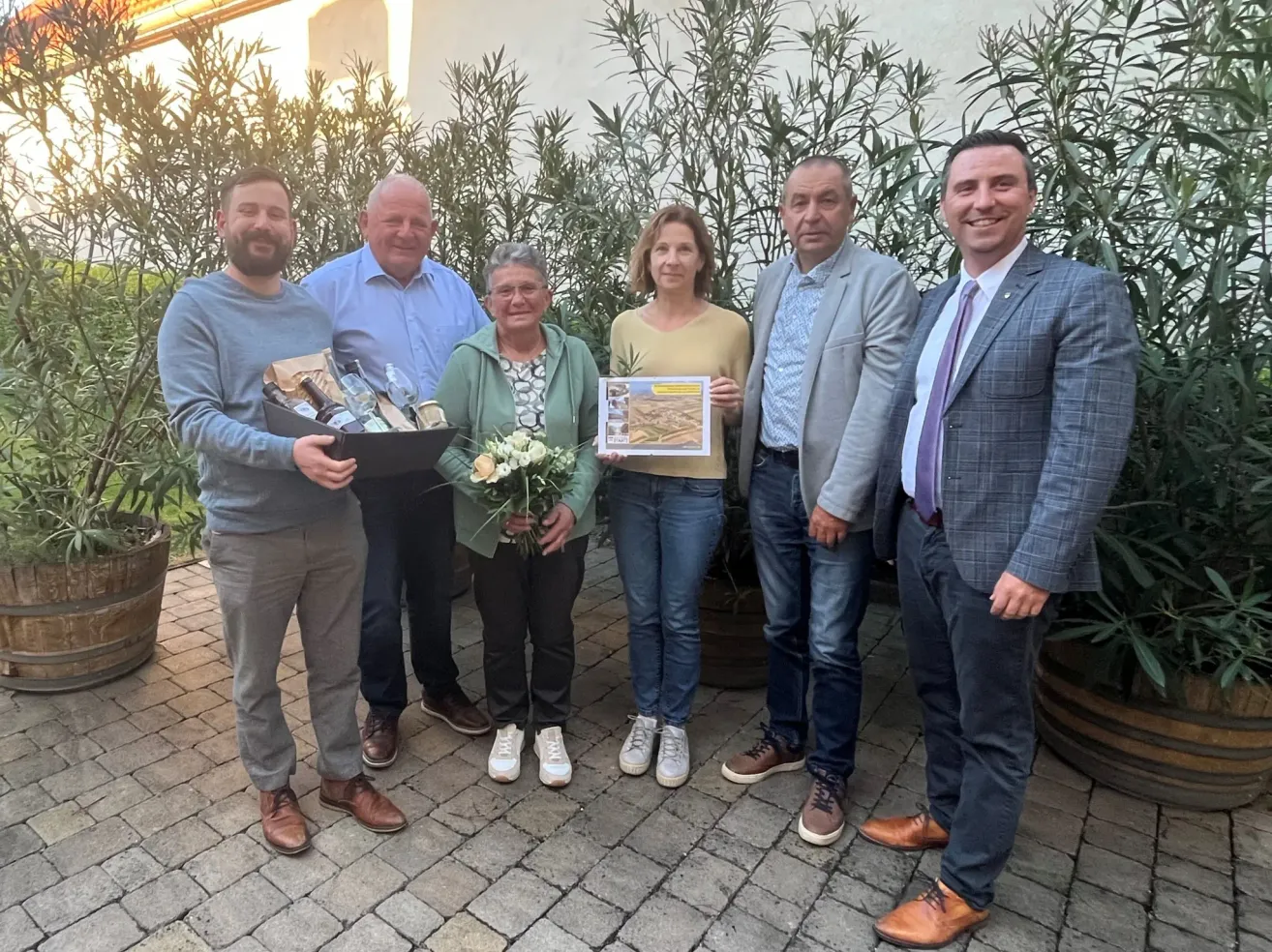Five individuals, including two women, stand outside holding a gift box and a certificate. They are surrounded by plants.