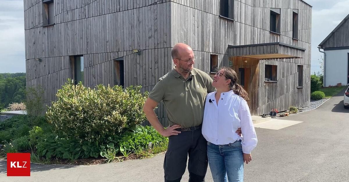 A man and woman stand in front of a modern wooden building with windows, greenery, and a small potted plant in the background.