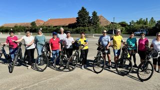 A group of cyclists, both men and women, standing in a parking lot, smiling, and posing for a photo. Some wear helmets, and all have bikes. In the background, there are houses and trees.