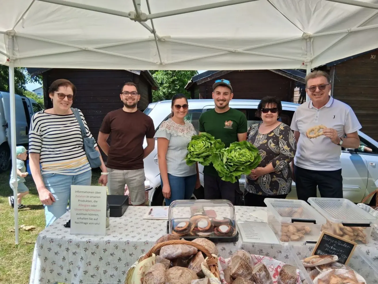 A group of people stand at a market stall under a tent, smiling and posing for a photo. They hold leafy greens, and the table is filled with various foods. A car is parked behind them.