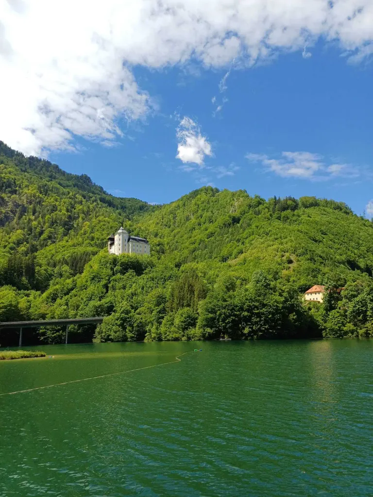 Ein Schloss auf einem grünen Hügel, umgeben von üppigem Grün. Unten spiegelt ein ruhiger See den klaren blauen Himmel wider.