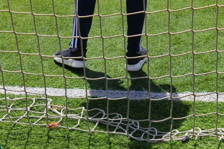 Eine Person in schwarzen Hosen und Turnschuhen steht hinter einem Fußballnetz. Das Netz ist an einer weißen Linie auf dem Grasfeld befestigt.