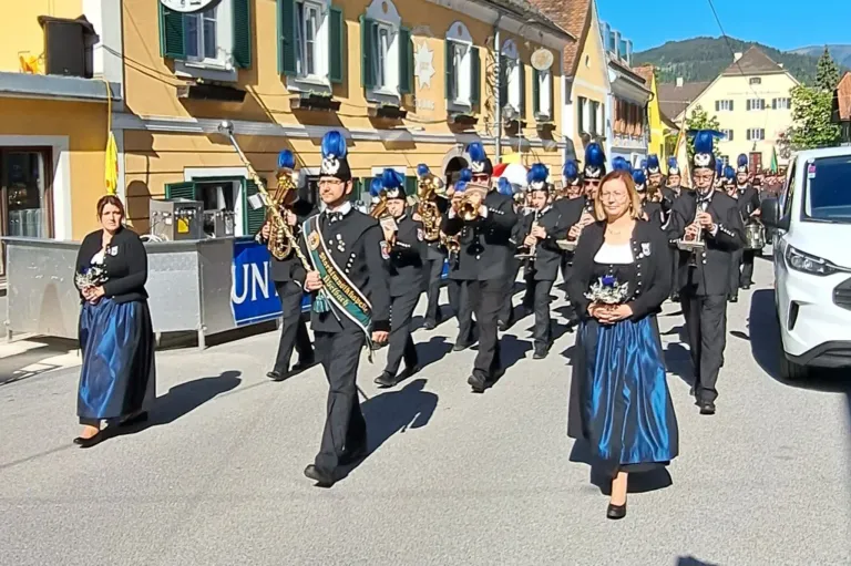 Ein Marschkapelle mit blauen Mützen und Uniformen, angeführt von einer Frau in einem blauen Kleid, marschiert vor einem Gebäude mit gelben Wänden.