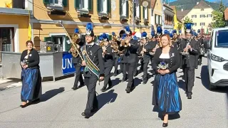 Ein Marschkapelle mit blauen Mützen und Uniformen, angeführt von einer Frau in einem blauen Kleid, marschiert vor einem Gebäude mit gelben Wänden.