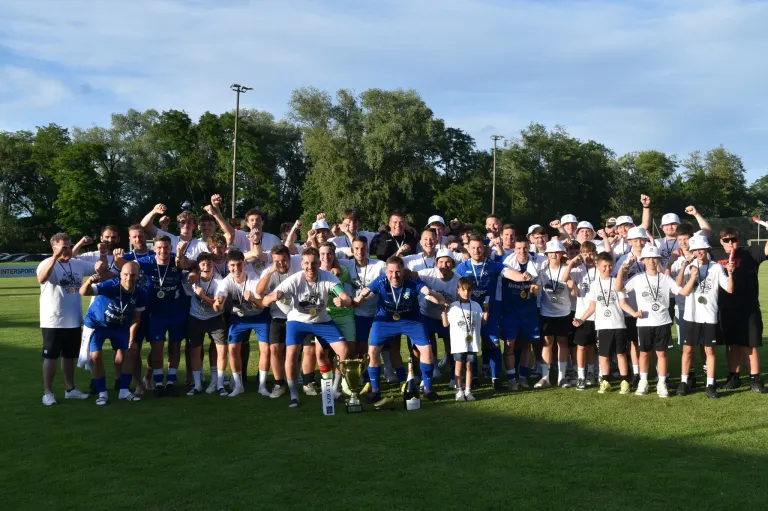 A group of people in blue and white uniforms celebrate on a grassy field with a trophy and medals. Trees and streetlights are in the background.