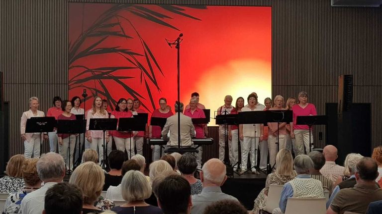 A choir of women and men in pink and white attire sings on stage. They have musical instruments in front of them and a large audience watches.