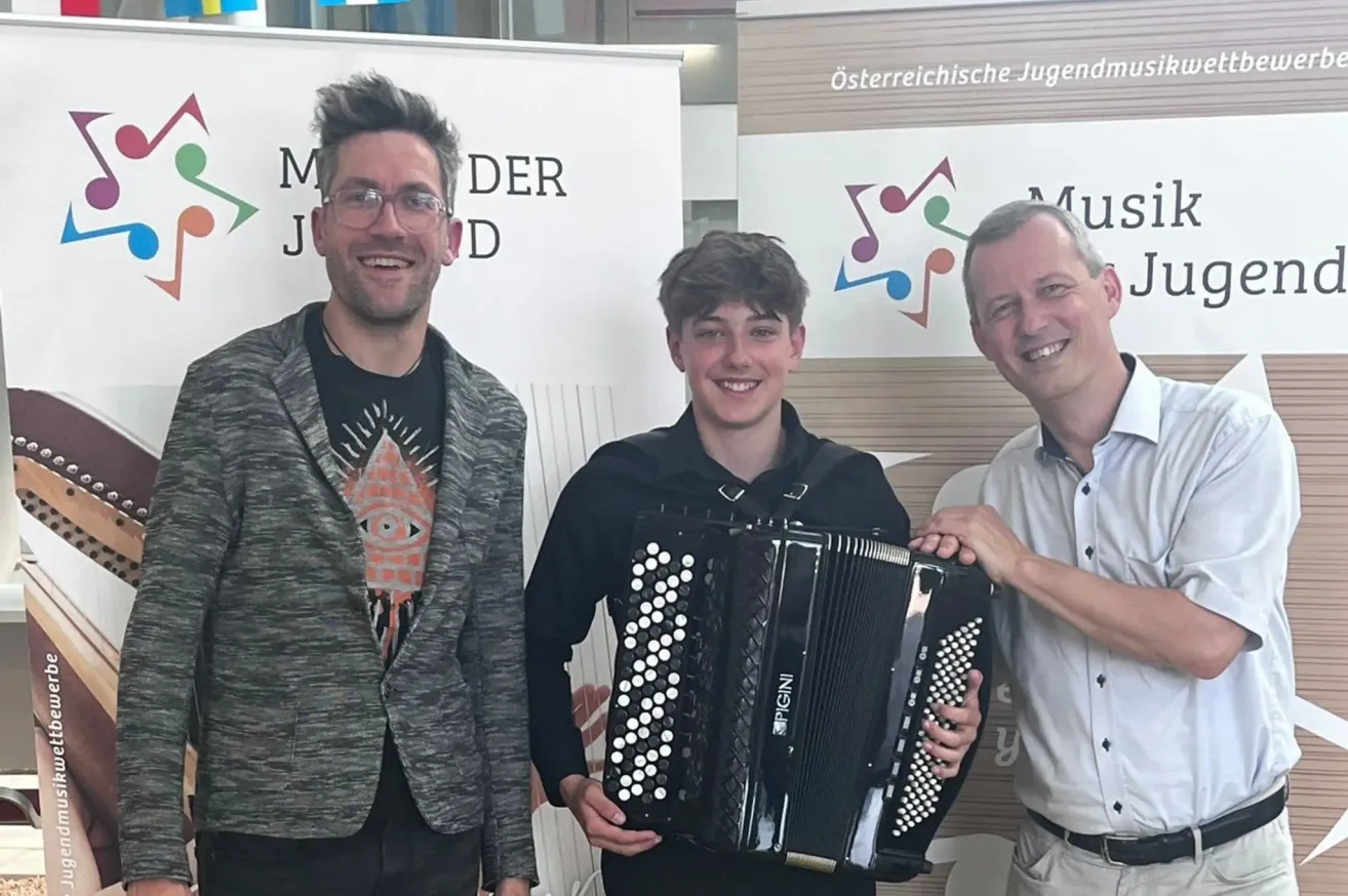 Three men pose for a photo. The man in the middle plays an accordion. The man on the right smiles. The background has a sign with music notes.