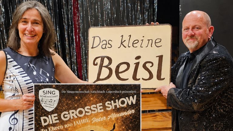 Ein Werbeplakat für 'Das kleine Beisl' wird von zwei Personen gezeigt. Das Plakat zeigt eine musikalische Aufführung und den Titel 'Die grosse Show'.