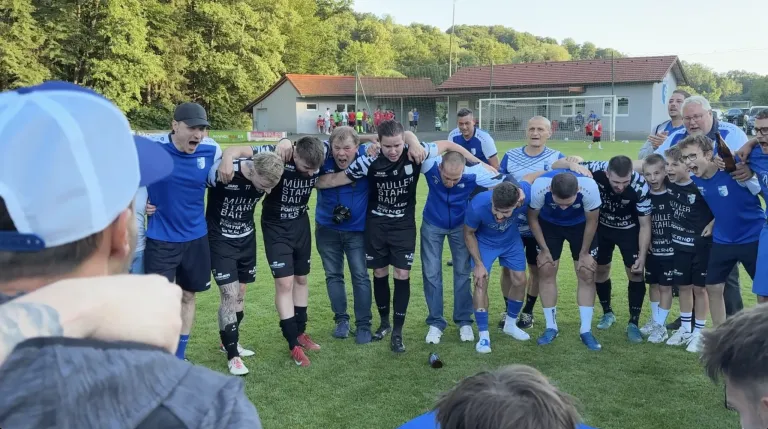 A soccer team in blue and white jerseys is huddled together on a grassy field, likely before a game. They are wearing shorts and sneakers. A bottle is on the ground near them.