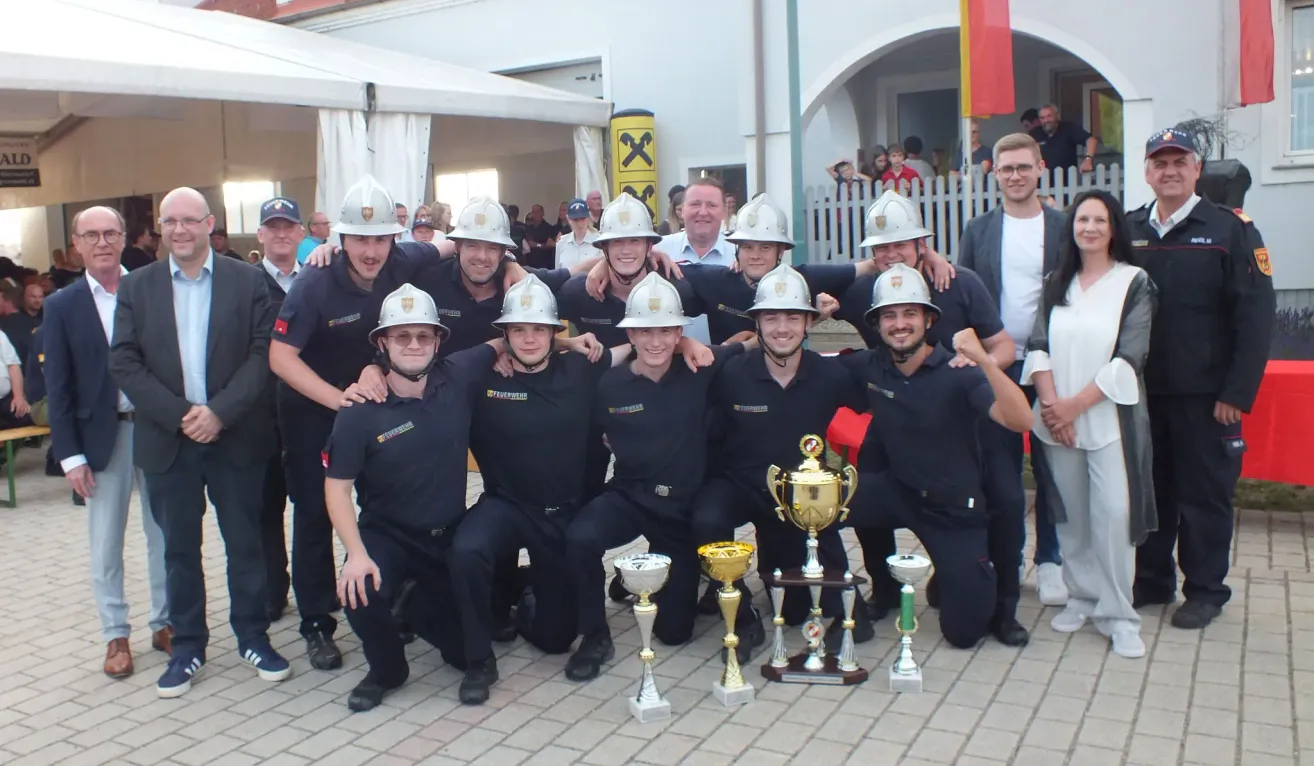 Eine Gruppe Feuerwehrleute in Uniform und Helmen posiert mit ihren Trophäen für ein Foto, während eine Menschenmenge im Hintergrund zuschaut.