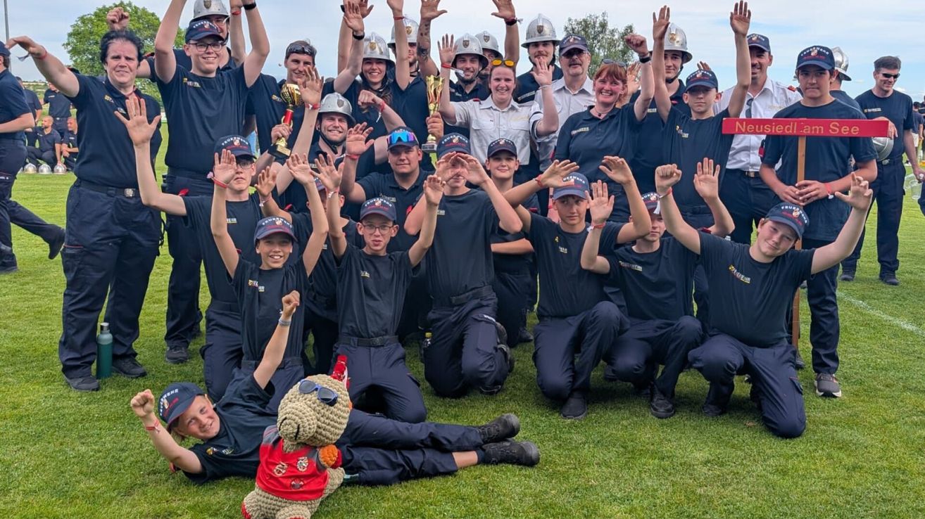 A group of firefighters wearing blue uniforms and helmets pose for a photo on a grassy field, some holding trophies and others raising their hands.