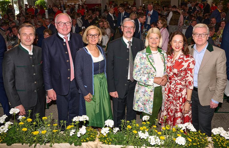 Sechs Personen stehen bei einem formellen Event mit Blumen vor ihnen. Dahinter versammelt sich eine Menschenmenge in einem großen Saal.